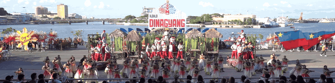 Cry of Jelicuon contingent performing in formation around bamboo hut set pieces on the riverside stage